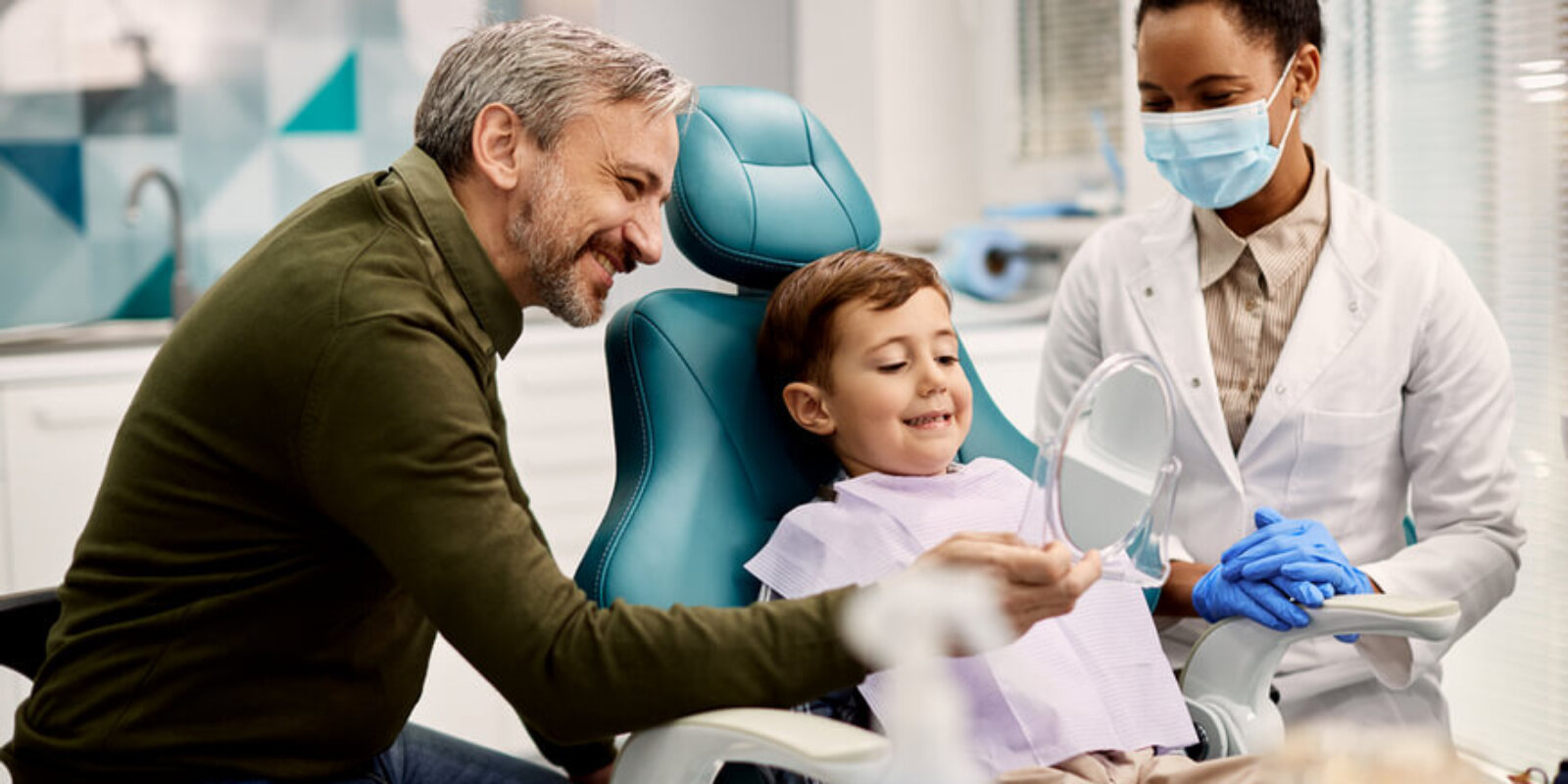Happy boy looking at his teeth in a mirror while being with his father at dentist's office.