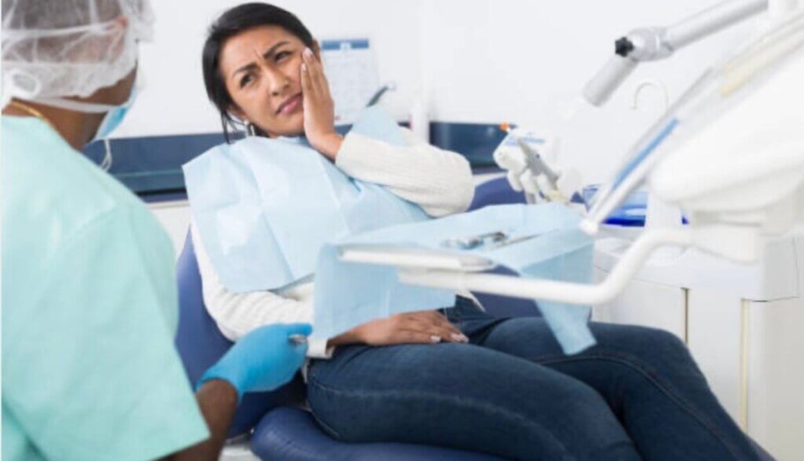 Woman sitting in a dental room while holding hand near face with discomfort and looking at a dentist.