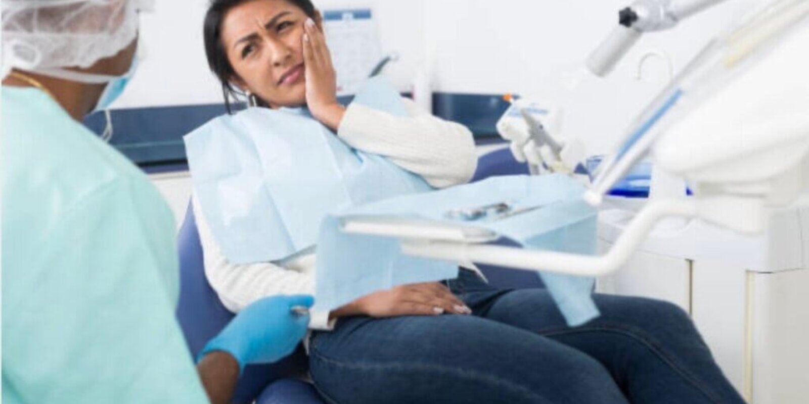 Woman sitting in a dental room while holding hand near face with discomfort and looking at a dentist.