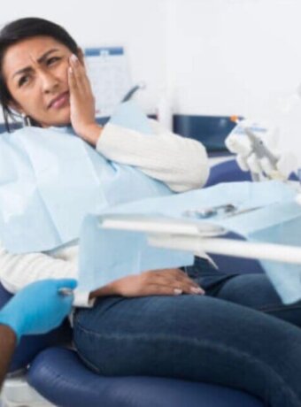 Woman sitting in a dental room while holding hand near face with discomfort and looking at a dentist.